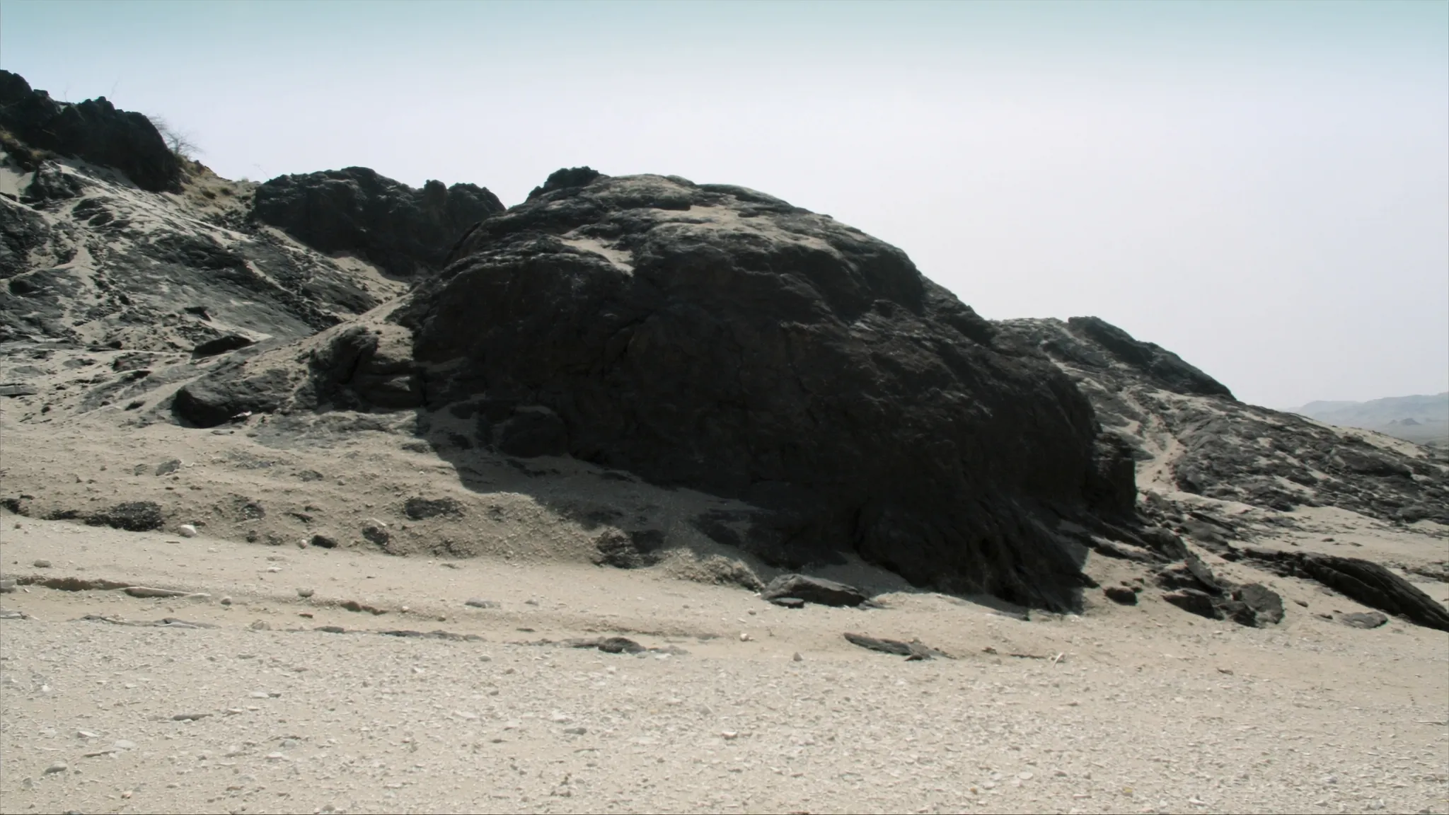 A weathered dark rock outcrop on a pale gravel plain under hazy light